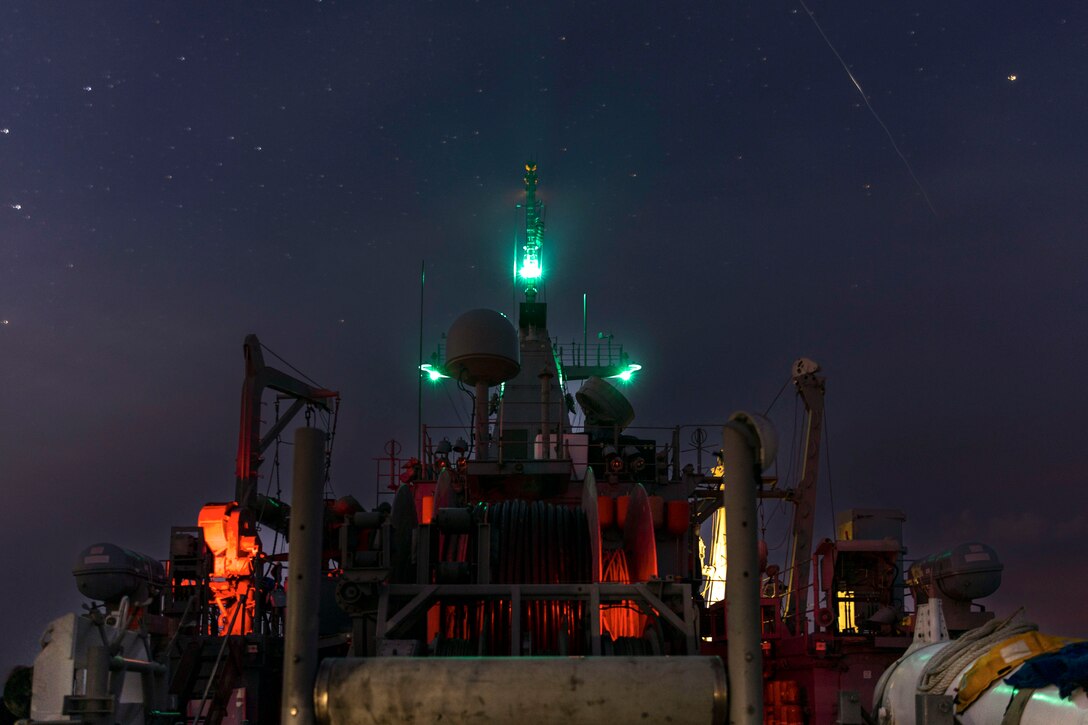 A ship sails under a starry sky in the dark illuminated by green, orange and yellow lights.