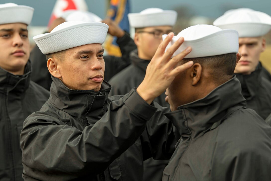 A sailor touches a sailor’s forehead as fellow sailors stand in formation in the background.