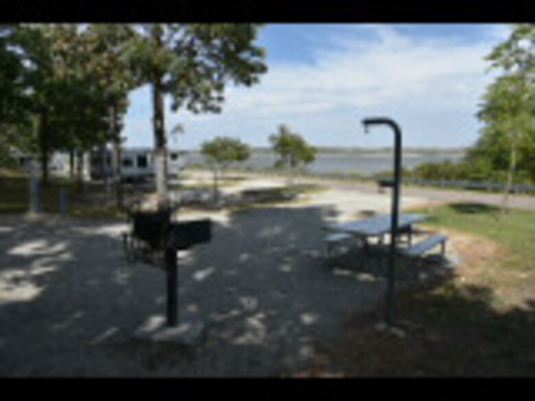 A campground can be seen with a grill and picnic table. Trees and the lake can be seen in the background.