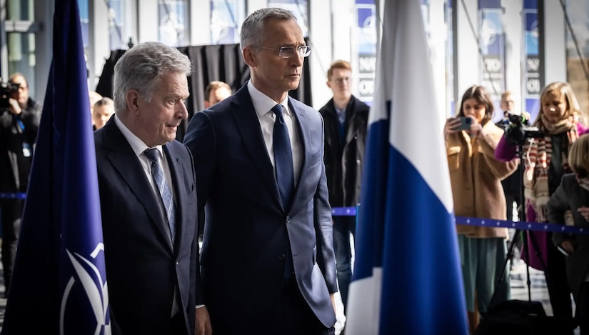 Two men in suits stand next to each other between two flags. Onlookers take photos.