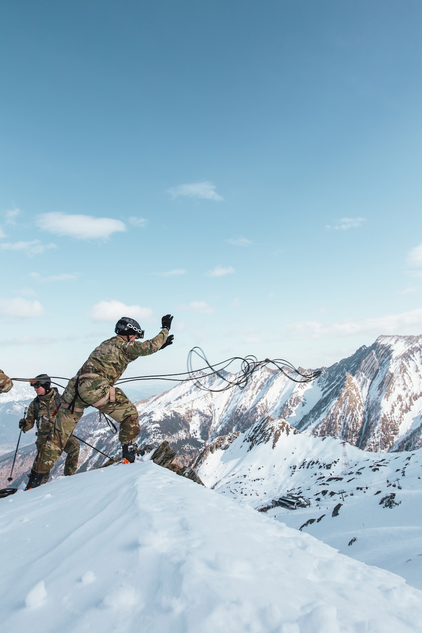A soldier tosses one end of a rope off a mountain ledge as others stand nearby.