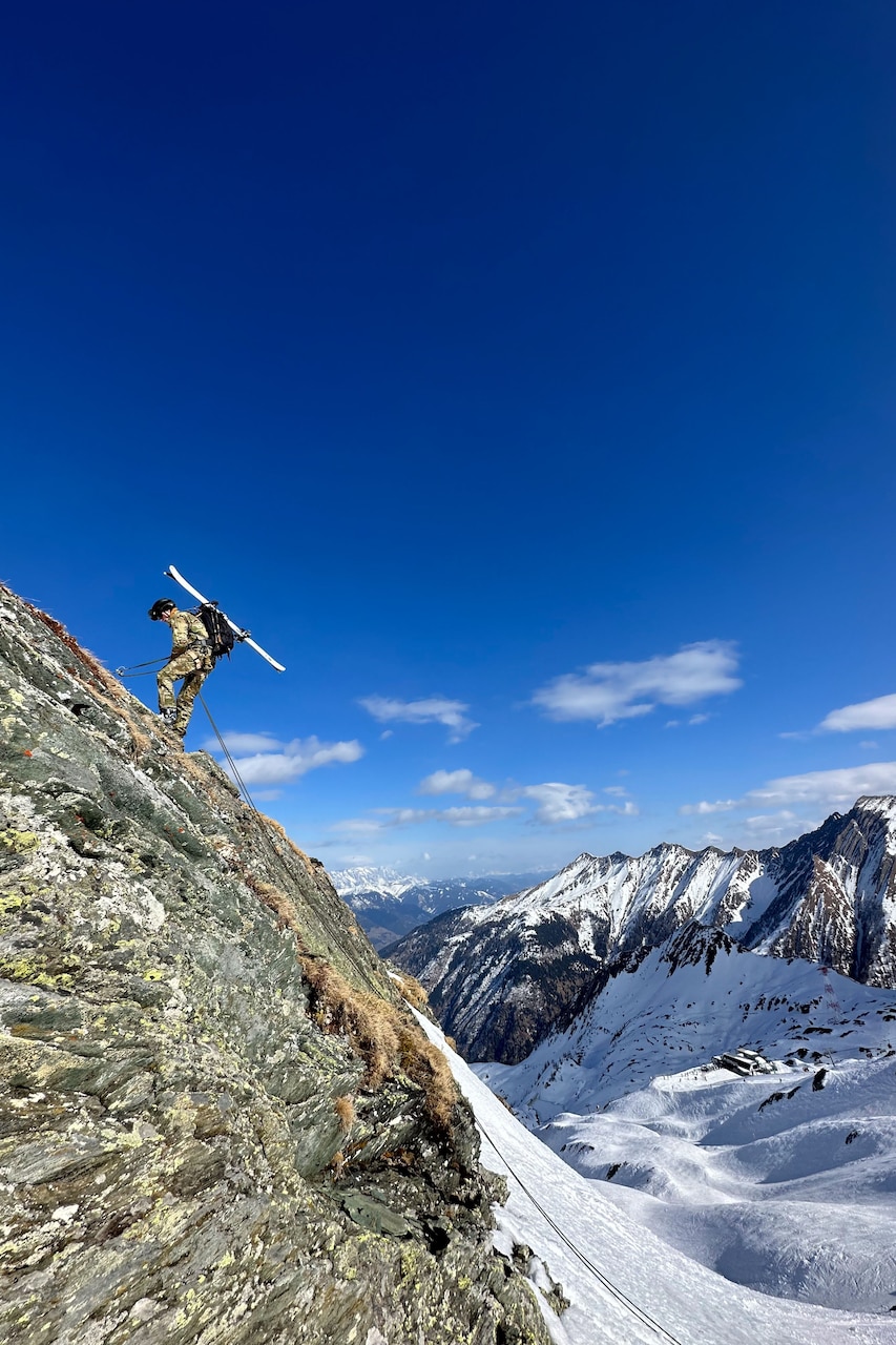 A soldier carrying skis attached to a backpack rappels on a rocky mountain face.