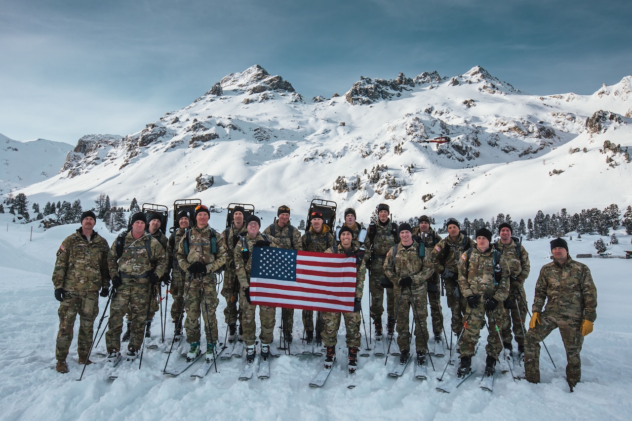 Soldiers on skis stand for a photo holding a U.S. flag, with snowy peaks in the background.