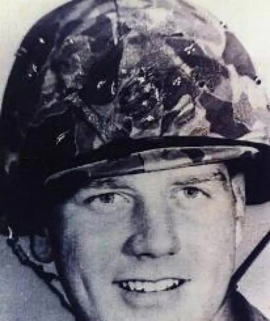 A man in a camouflage combat helmet smiles for a photo.