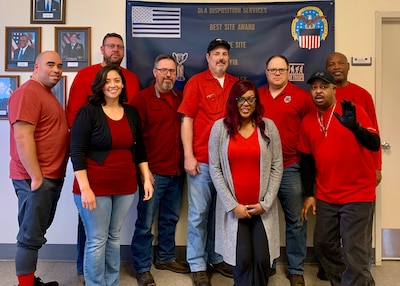 Nine people stand together each wearing a RED shirt. All are smiling and one person is waving to the camera.