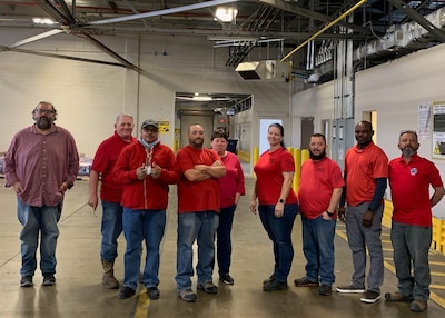 Nine adults all wearing RED shirts stand next to each other in a warehouse where they work.