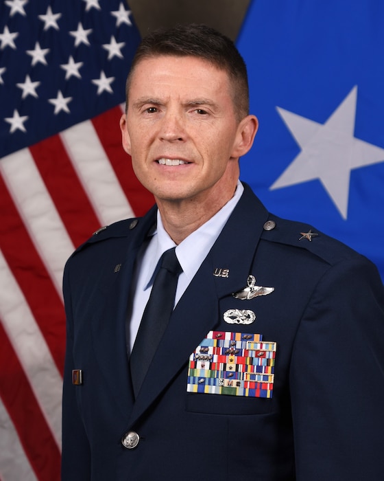 Service member smiles for official portrait in front of U.S. Flag and one star flag.