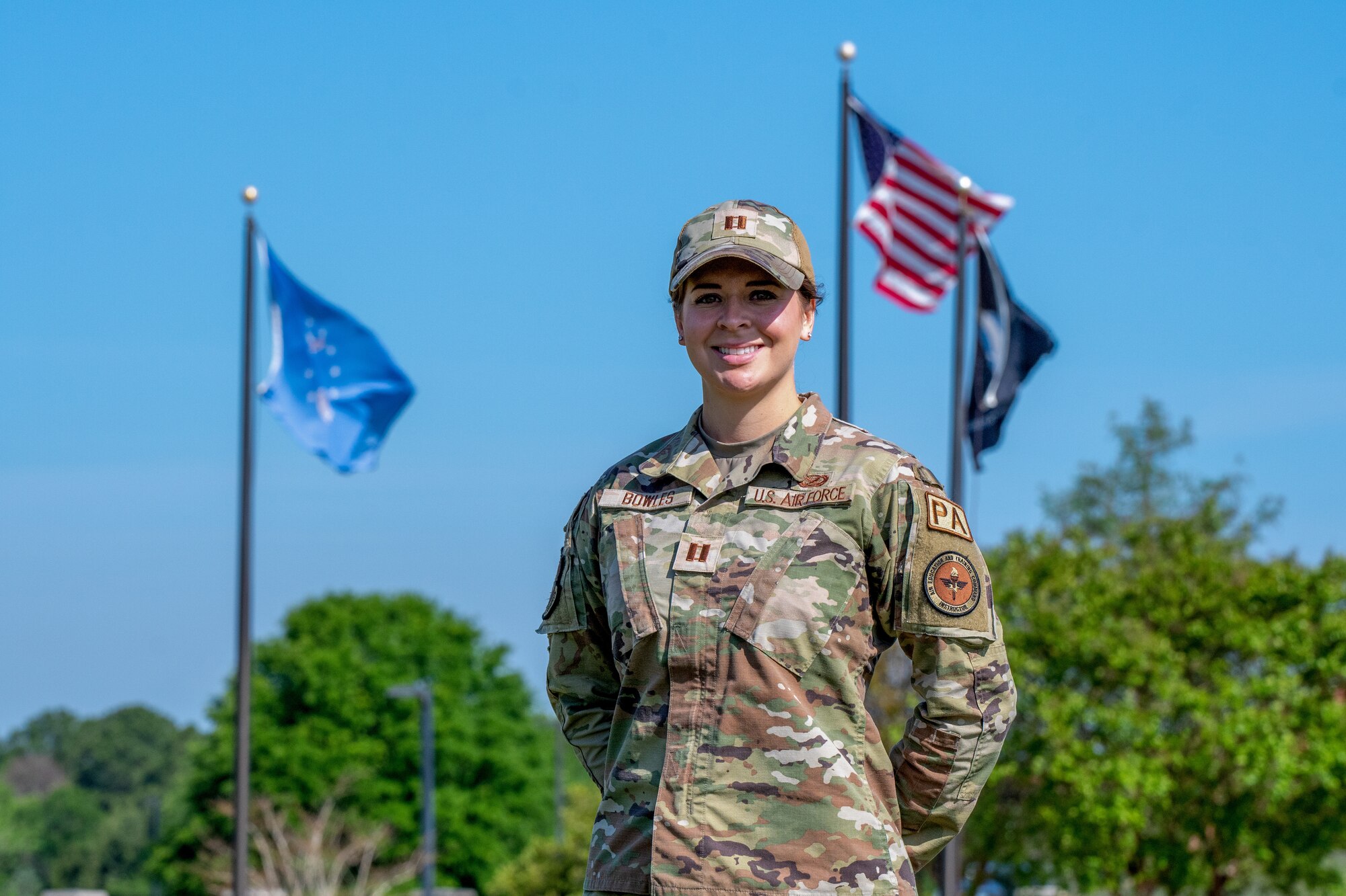 Capt. Abigail Bowles stands in her OCP uniform in front of flag poles and tress, smiling at parade rest.