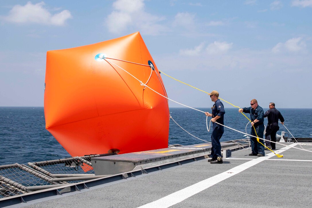 Sailors lower a large red inflatable target into water from the deck of a ship.