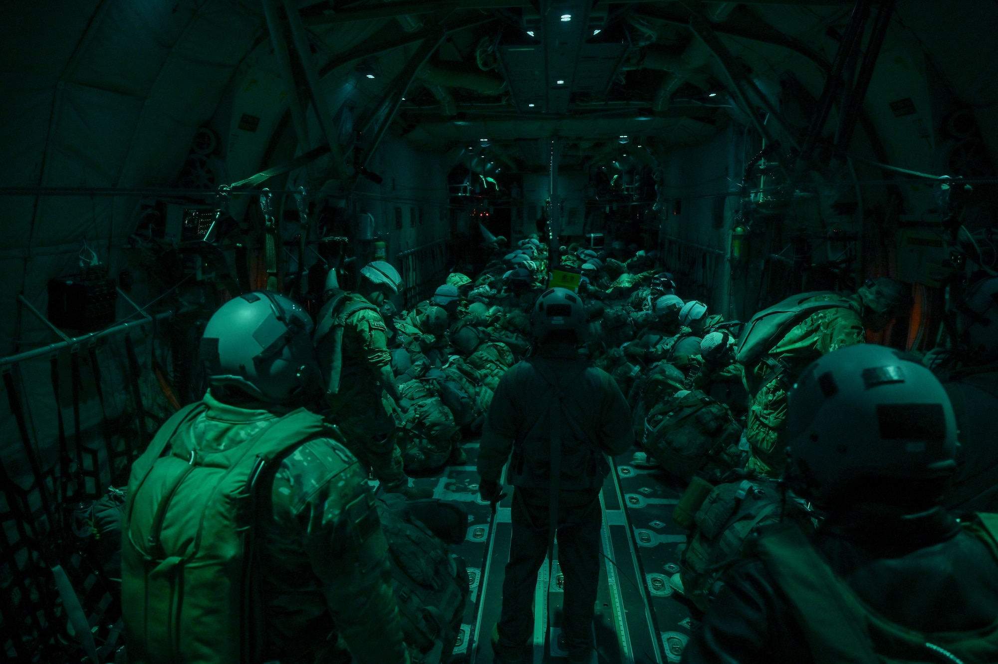 Three men in flight suits standing facing the men sitting down in a c130j aircraft. the room is dark green.