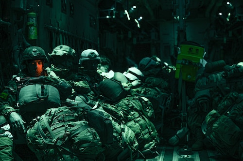 Paratroopers with combat gear sit resting in a dark room that is lit with green lights. One mans face is lit with an orange hue due to the light coming through the window of the aircraft.