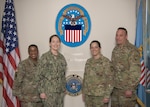 A group of four U.S. military service members pose for a photo in front of the Defense Logistics Agency's logo on a wall.