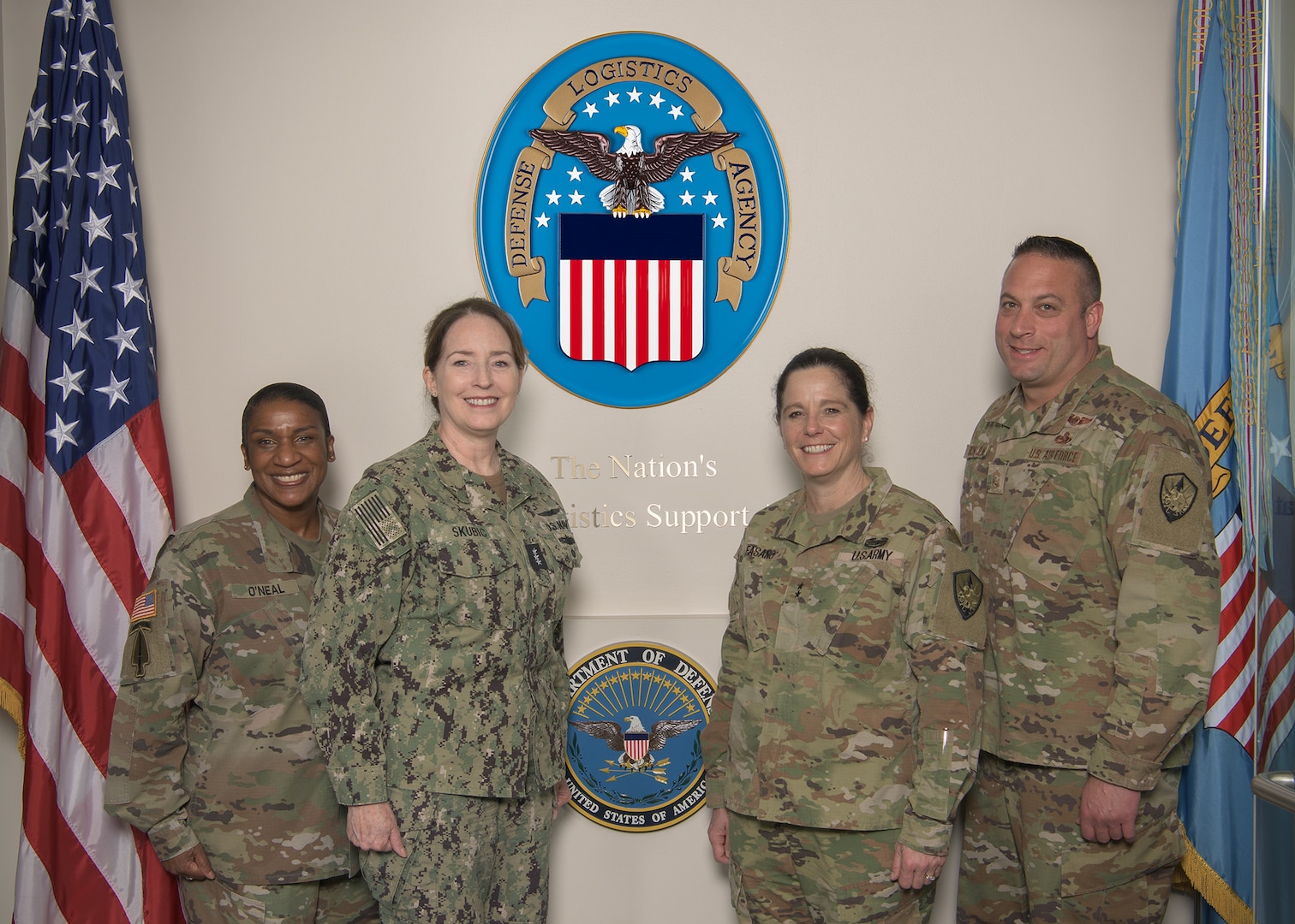 A group of four U.S. military service members pose for a photo in front of the Defense Logistics Agency's logo on a wall.