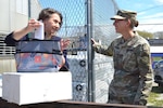 Mid-close up photo of two white women standing outside on a sunny day. Woman on left has brown curly hair pulled back and woman on right is wearing an army camo uniform and hat, with blond hair in low ponytail. Woman on left is handing woman on right a package of seeds while talking. both are smiling and look happy.