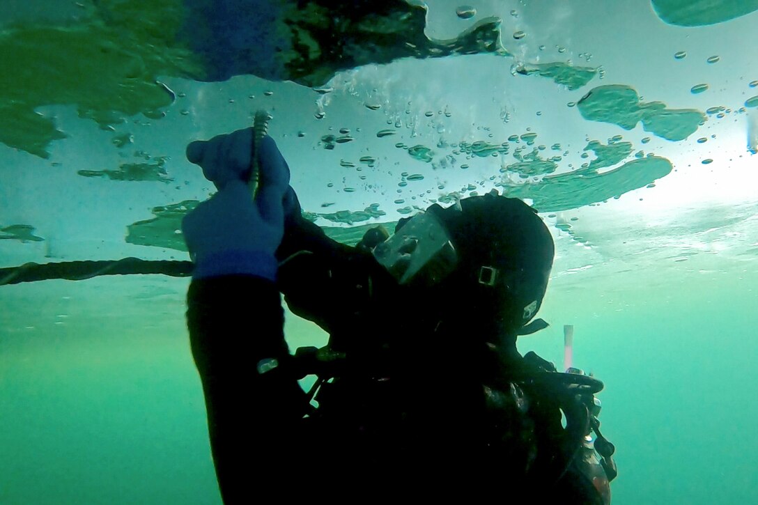 A diver swimming underwater inserts a screw into ice in a frozen lake.
