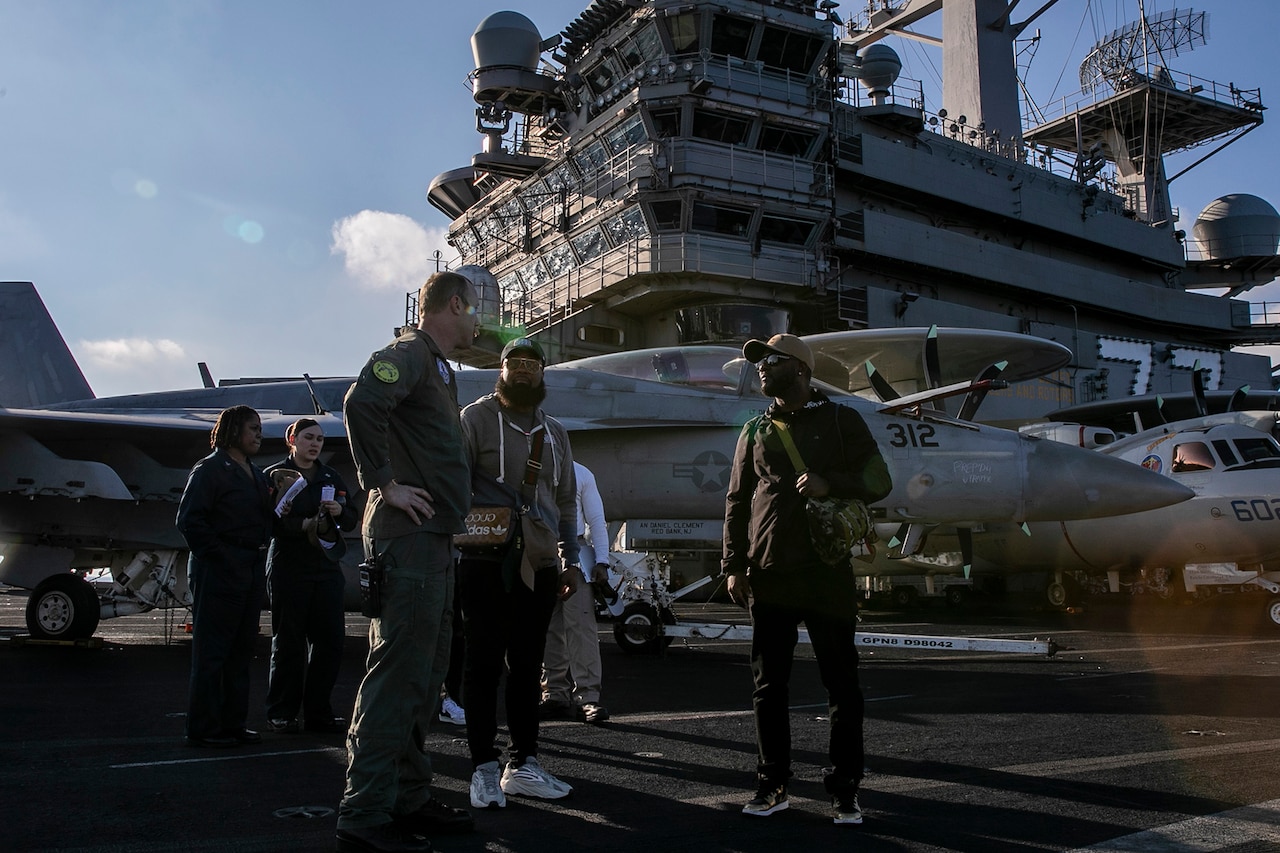 Three men talk on the deck of a large ship. A few others stand behind them near a fighter jet.