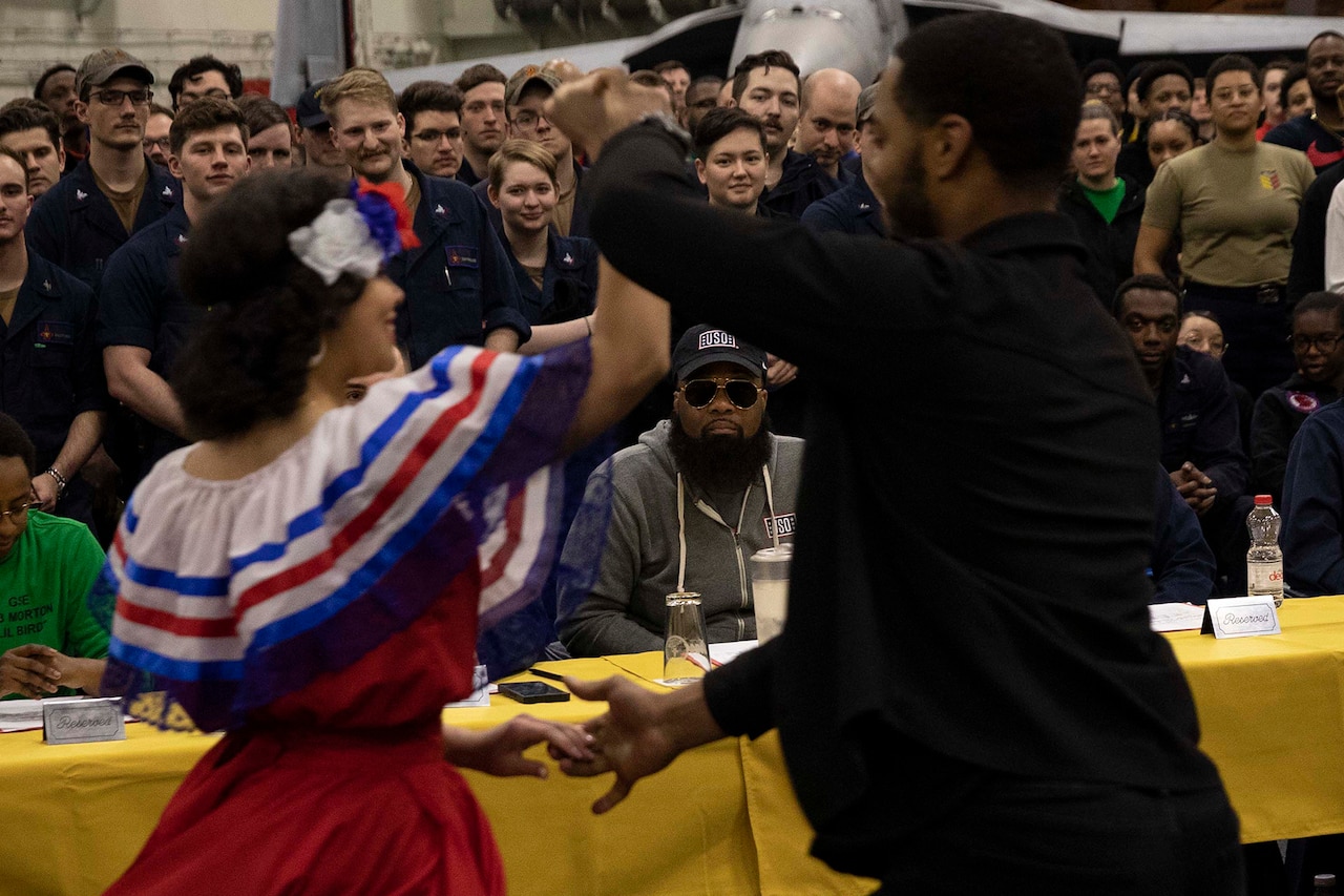 A man at a table watches a couple dance. A large group stands behind the man at the table.