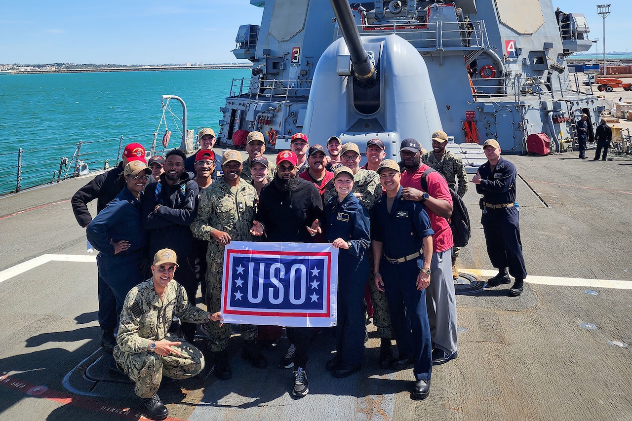 Several people pose with a USO sign on the deck of a large ship. A massive gun sits in the background.