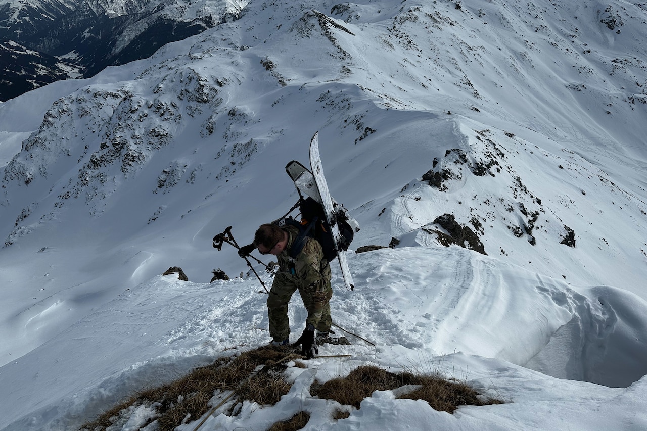 A soldier wearing skis on his back climbs a snowy ridge.