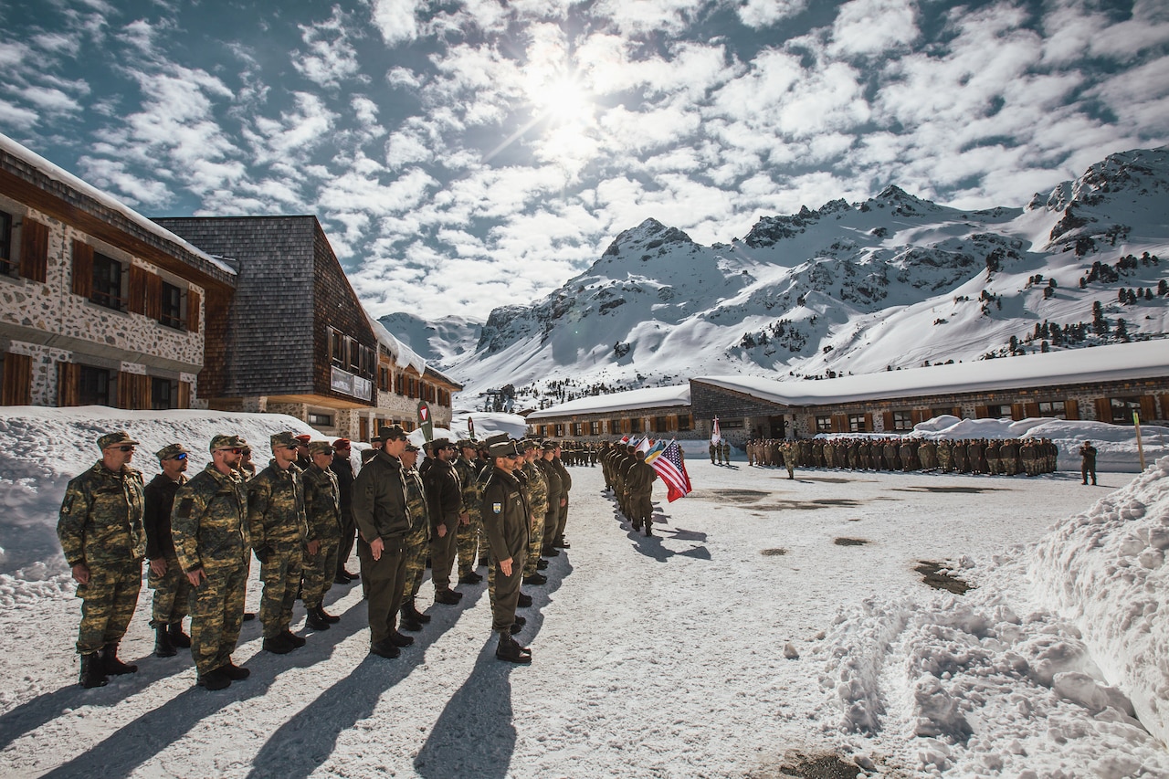 Multinational service members stand in formation outside buildings set in the mountains.