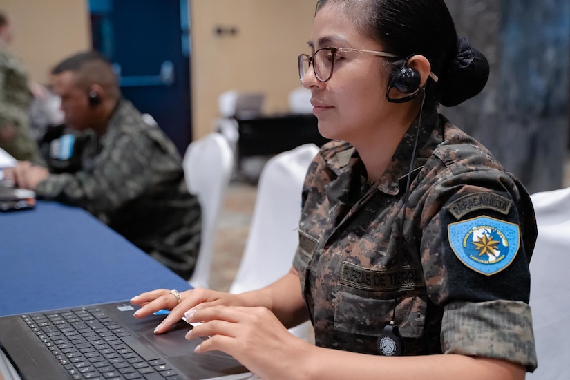 A female soldier works on a laptop, as does a soldier in the background.
