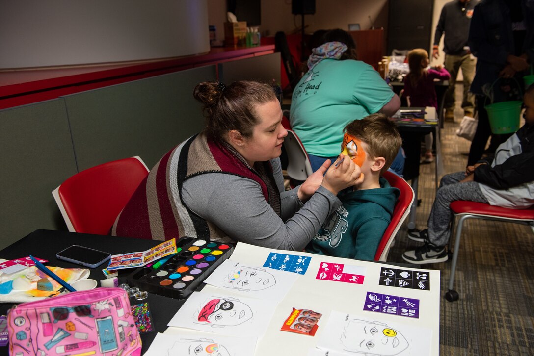 A woman in a grey shirt paints a tiger on the face of a brown haired boy in a green hoodie.