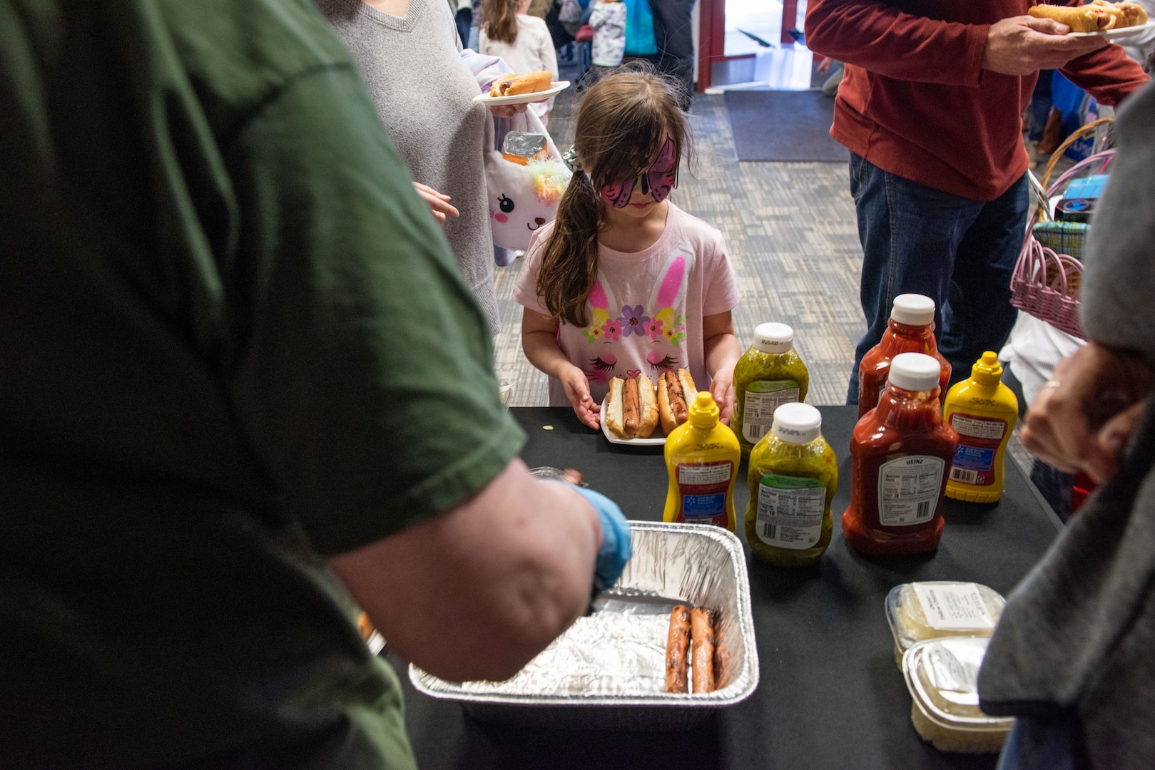 A girl with a butterfly painted on her face grabs two hot dogs from a table. She wears a pink shirt.