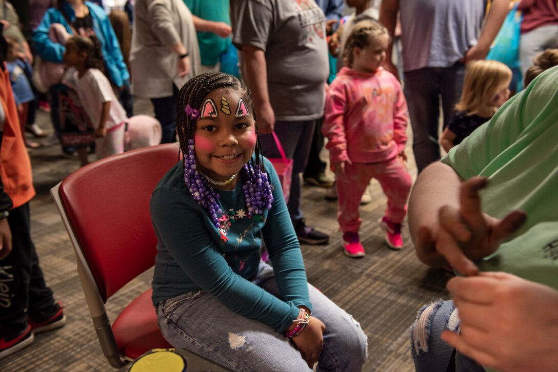 A small dark skinned girl has her face painted. She wears a green shirt and jeans.