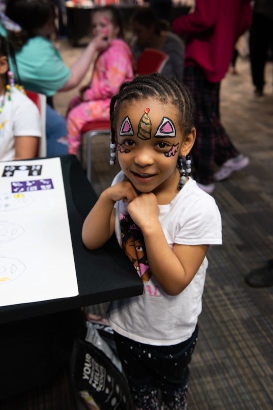 A small dark skinned child in a white shirt shows off her painted face.