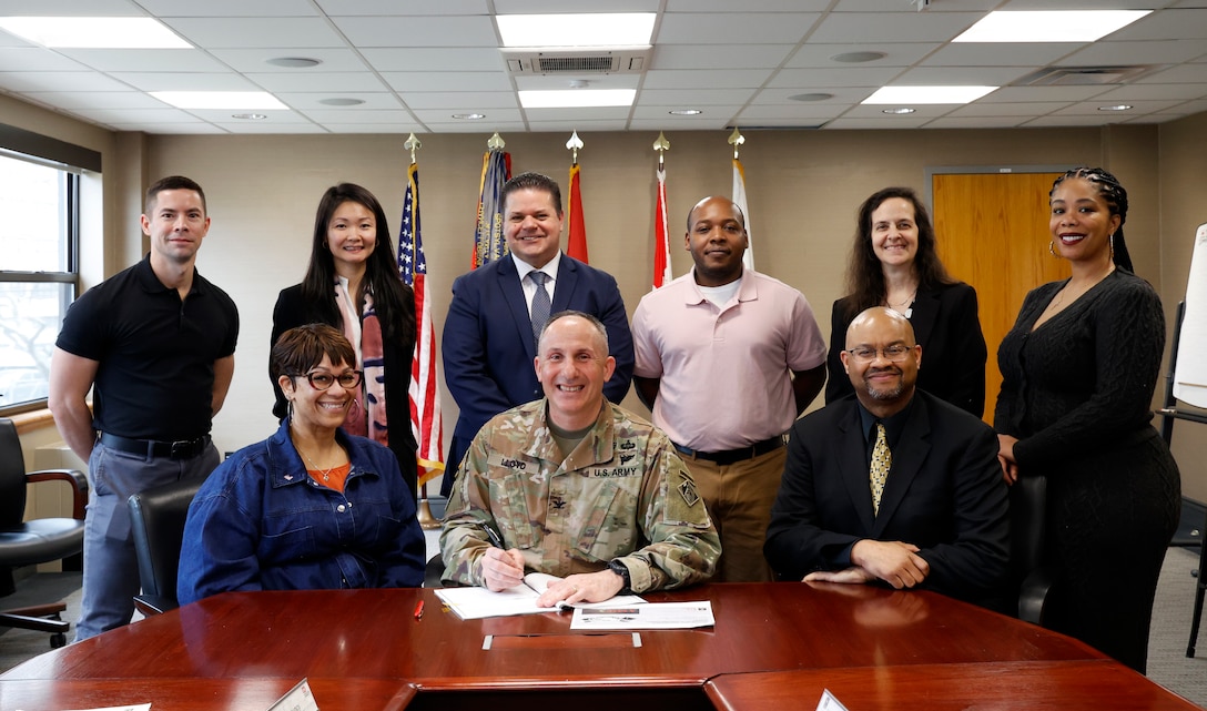 Nine people gather in an office to sign a document.