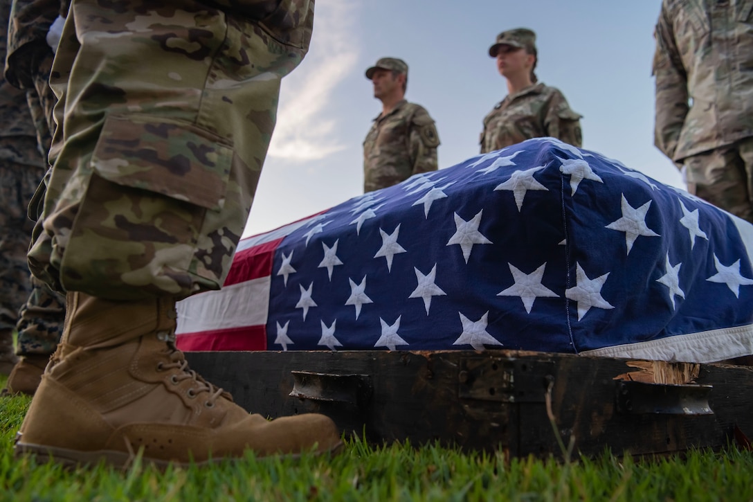 Service members stand next to a casket with an American flag draped over it.