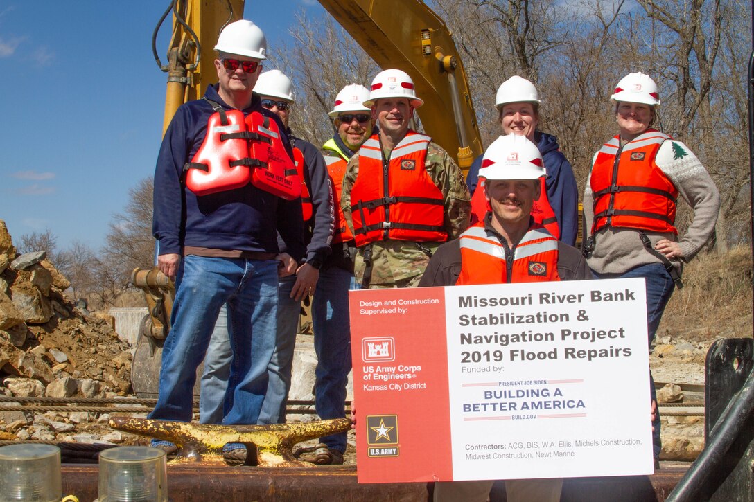 Seven people pose in front of a crane and a large pile of rocks on a barge. The person in the front is holding a sign that says "Missouri River Bank Stabilization and Navigation Project 2019 Flood Repairs"