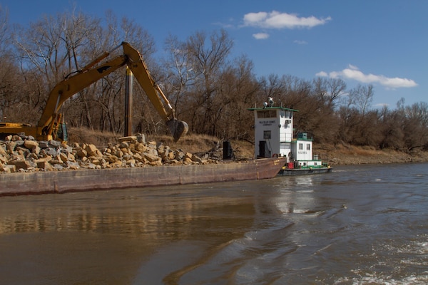 A barge is on the river with a crane placing rock on the river bank. Trees are in the background.