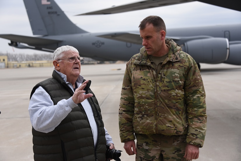 Two men stand near a military aircraft.