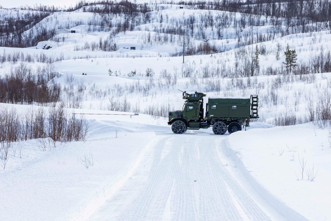 A military vehicle stops in front of a barrier arm at the juncture of two snow-covered roads with hilly terrain in the background.