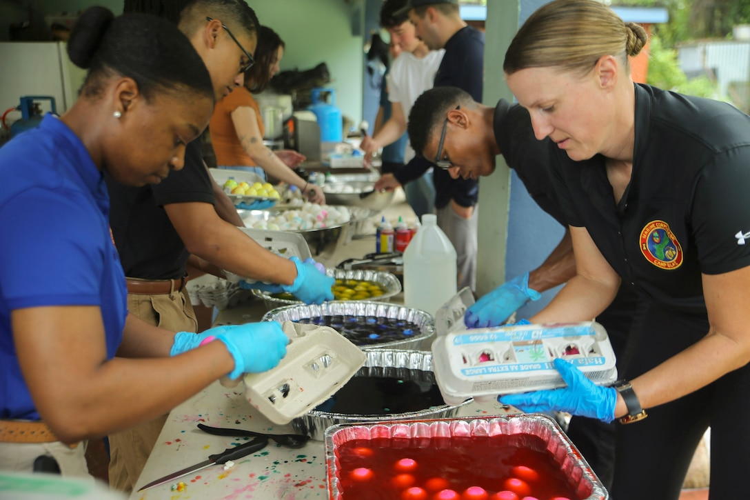 Marines standing on opposite sides of a table place eggs into aluminum bowls of red, blue and yellow dye.