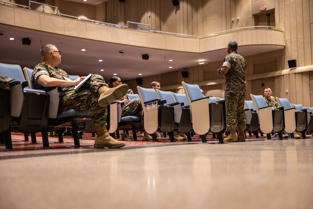 A Marine speaking in front of an auditorium with a few other Marines seated in front of him.
