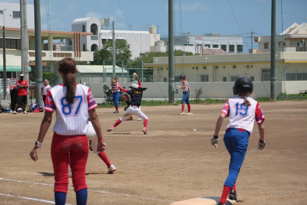 Women in softball uniforms playing on a diamond with the pitcher throwing a ball from the mound.