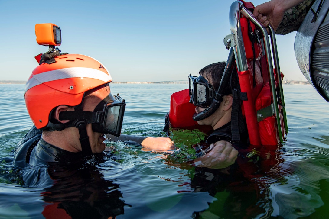 A sailor in body of water holds onto a fellow sailor attached to a stretcher next to an inflatable boat.