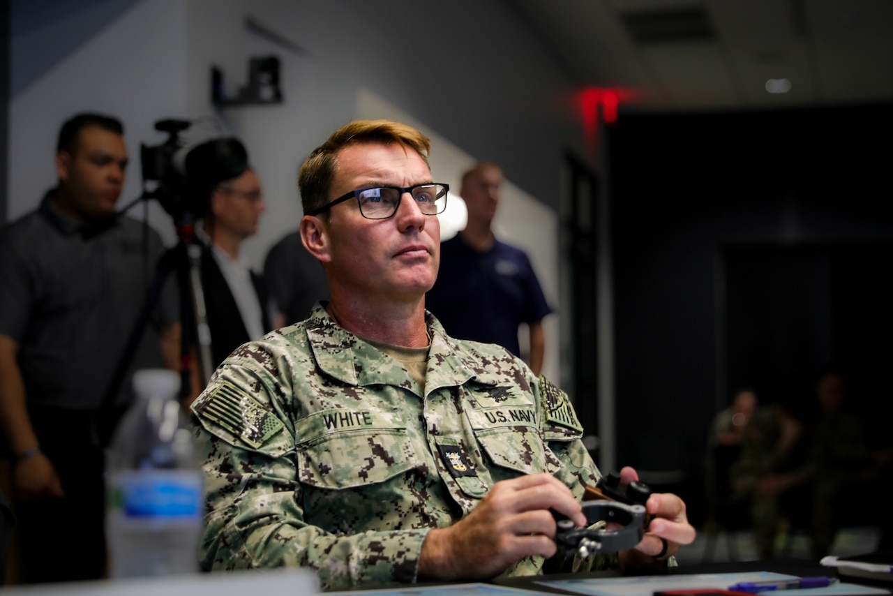 One man sitting at a table in uniform holds a 3D printed device.