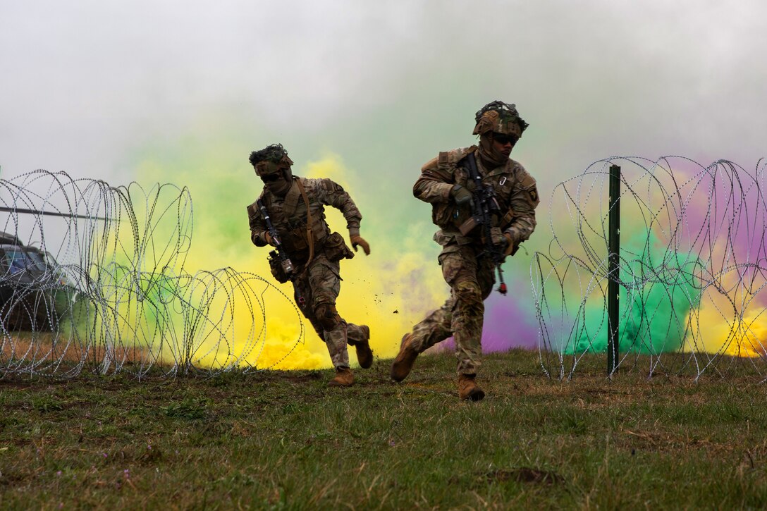 Soldiers run through a field of multi-colored smoke and barred wire obstacles.