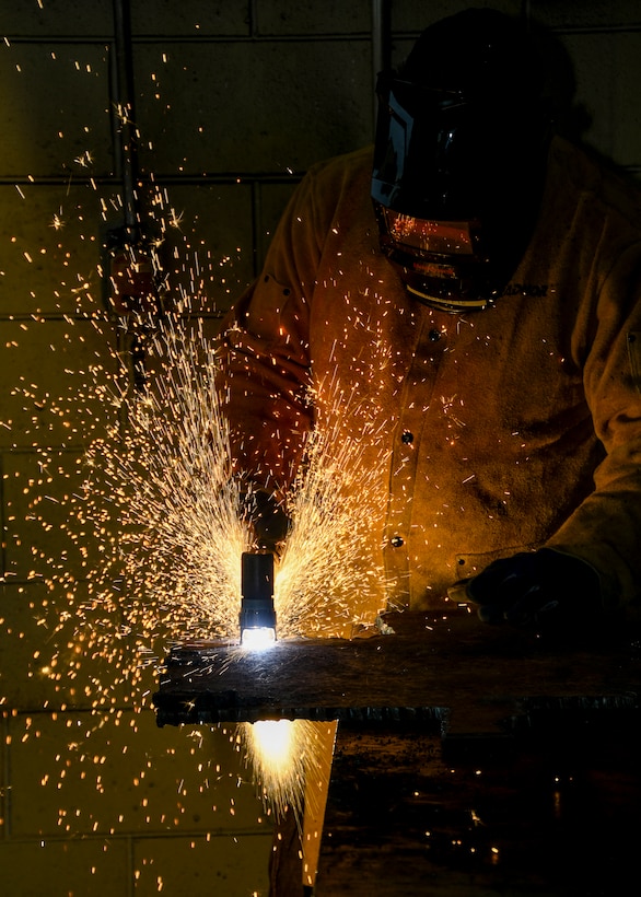 A worker in protective gear uses a plasma cutter emitting a spray of bright sparks against a darkened background