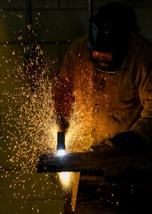 A worker in protective gear uses a plasma cutter emitting a spray of bright sparks against a darkened background