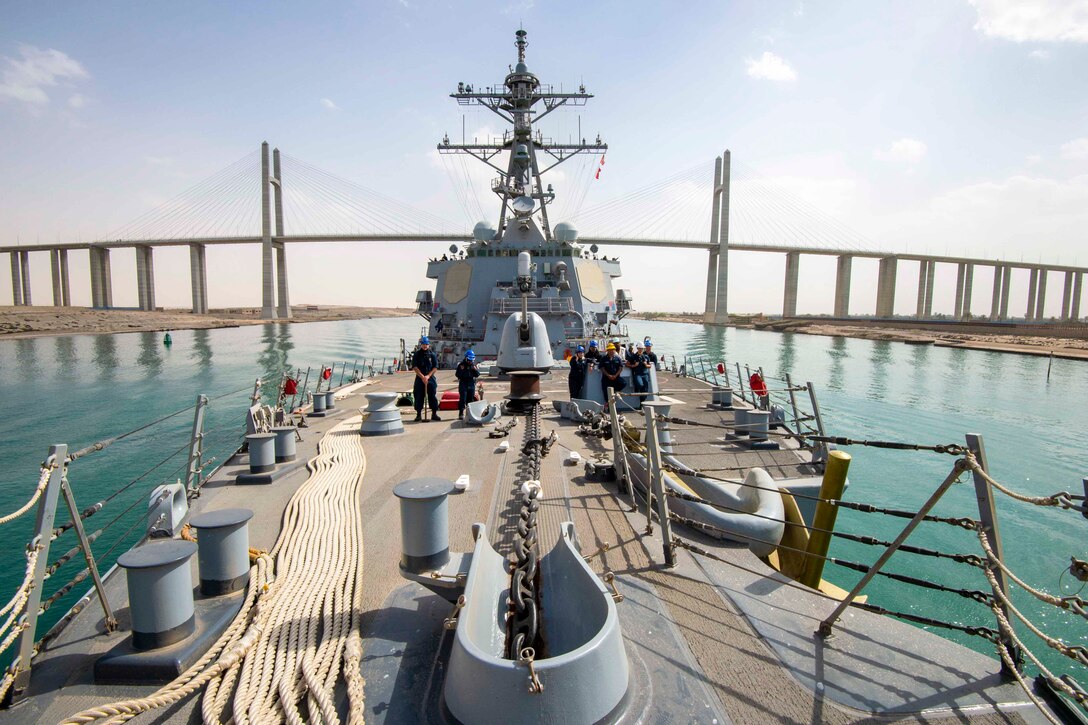 Sailors stand on the deck of a ship as it travels through a body of water toward a bridge.