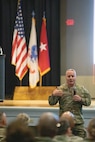 A Soldier standing with a microphone speaks to Soldiers in an auditorium who are sitting.