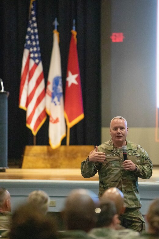 A Soldier standing with a microphone speaks to Soldiers in an auditorium who are sitting.