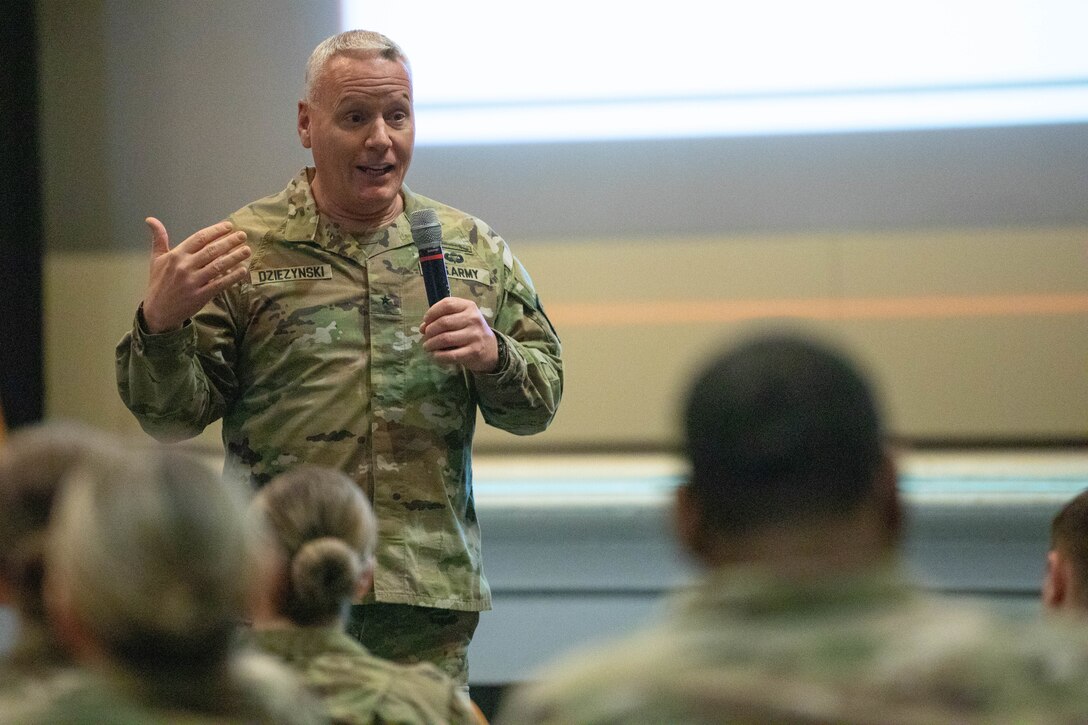 Standing Soldier with microphone speaking to Soldiers sitting down in an auditorium.