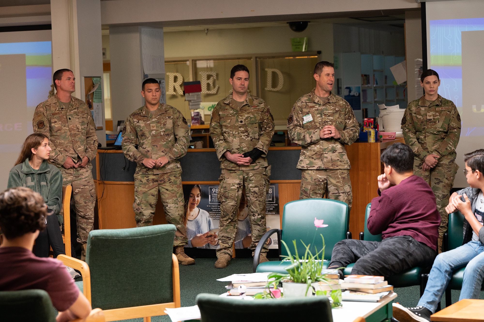 Airmen stand in a library and speak to teens at a high school