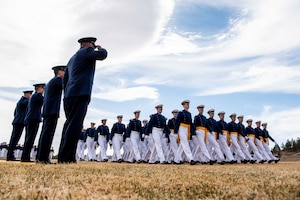 Cadets march in formation as airmen stand and salute.