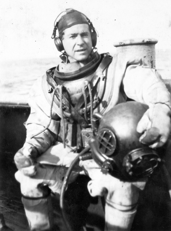 A man in an early 1900’s-era dive suit sits for a photo while holding a heavy metal helmet.